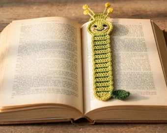 May include: A crochet bookmark shaped like a green caterpillar with a smiling face, pink cheeks, and two yellow antennae, resting inside an open book with printed text on the pages. The caterpillar has a segmented body and a small dark green leaf.