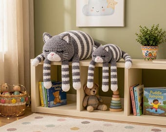 May include: Two handmade crochet cat plushies in varying sizes, featuring grey and white horizontal stripes, white paws, and white muzzles with pink noses. The cats have closed eyes with embroidered eyelashes and long, dangling limbs, resting on a shelf.