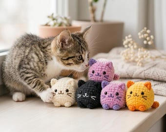 May include: A tabby kitten interacts with five small, round, crocheted cat plushies. The plushies are colored cream, black, light purple, lavender, and orange. Each features embroidered eyes, a small nose, and whiskers, with tiny ears.