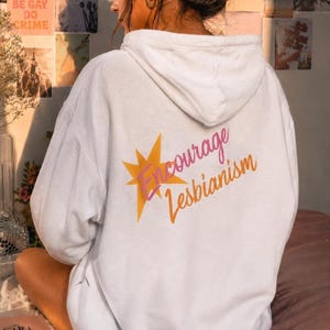 May include: A white hooded sweatshirt featuring a large yellow star graphic on the back. Overlaid on the star is the text "Encourage Lesbianism" written in a pink and orange cursive script. The hoodie has long sleeves and a hood with drawstrings.