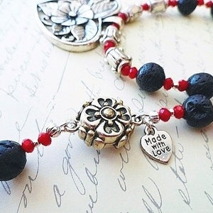 May include: A silver necklace with black lava rock beads and red glass beads. The necklace has a silver charm with a flower design and a silver heart charm with the text "Made with Love".