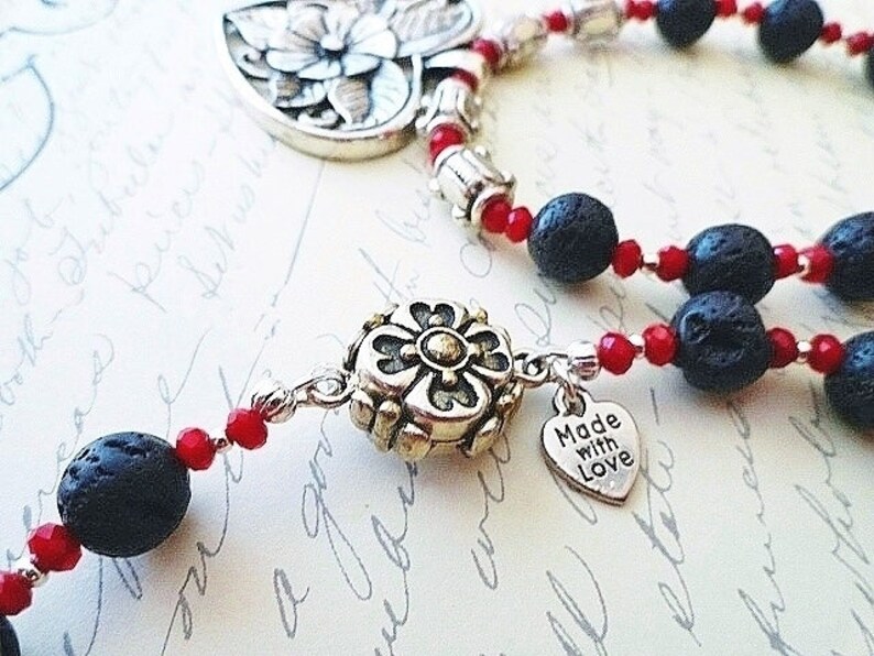 May include: A silver necklace with black lava rock beads and red glass beads. The necklace has a silver charm with a flower design and a silver heart charm with the text "Made with Love".