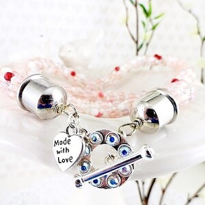 May include: A delicate pink beaded bracelet with silver-toned metal accents. The bracelet features a toggle clasp adorned with blue crystals and a heart charm that reads "Made with Love."