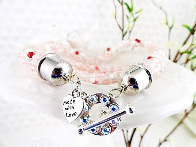 May include: A delicate pink beaded bracelet with silver-toned metal accents. The bracelet features a toggle clasp adorned with blue crystals and a heart charm that reads "Made with Love."