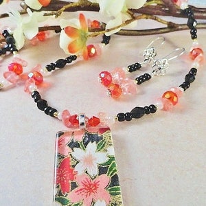 May include: A delicate necklace with a rectangular pendant featuring a floral design. The necklace is made with black beads, pink and red glass beads, and a silver chain. The pendant is made of glass and has a floral pattern with pink and red flowers on a black background.