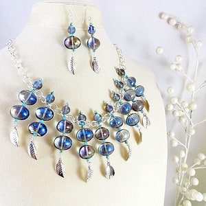 May include: A necklace and earring set featuring iridescent blue and silver beads. The necklace has a silver chain and is adorned with multiple beads and silver leaf charms. The earrings mirror the necklace design, creating a cohesive jewelry set.