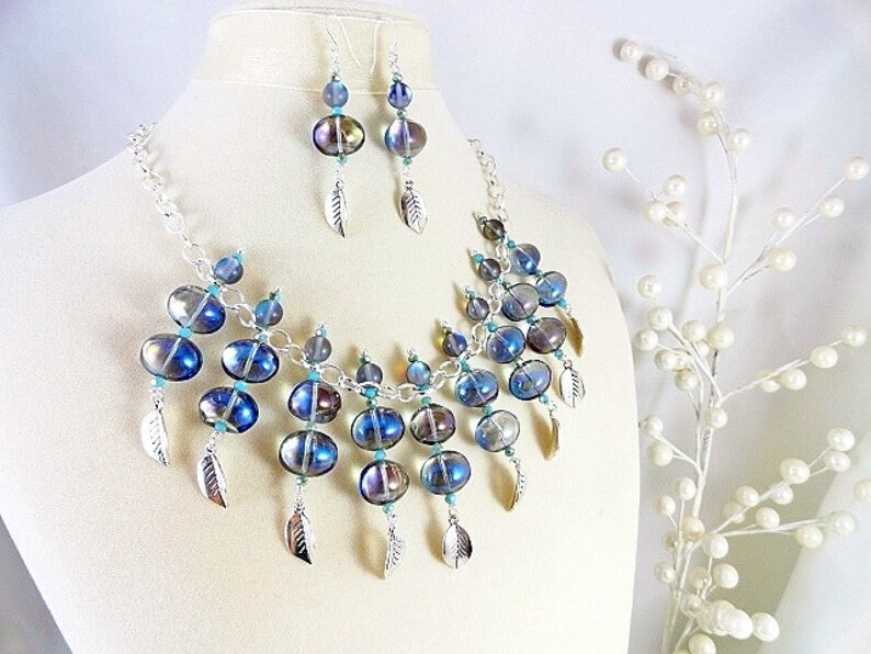 May include: A necklace and earring set featuring iridescent blue and silver beads. The necklace has a silver chain and is adorned with multiple beads and silver leaf charms. The earrings mirror the necklace design, creating a cohesive jewelry set.