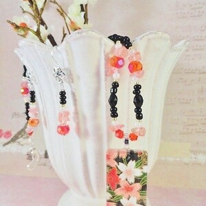 May include: A black and pink beaded necklace with a rectangular pendant featuring a floral design. The pendant is made of paper and has a gold background with pink and red flowers. The necklace is displayed on a white ceramic vase with a floral design.