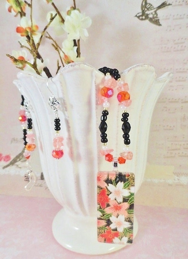 May include: A black and pink beaded necklace with a rectangular pendant featuring a floral design. The pendant is made of paper and has a gold background with pink and red flowers. The necklace is displayed on a white ceramic vase with a floral design.