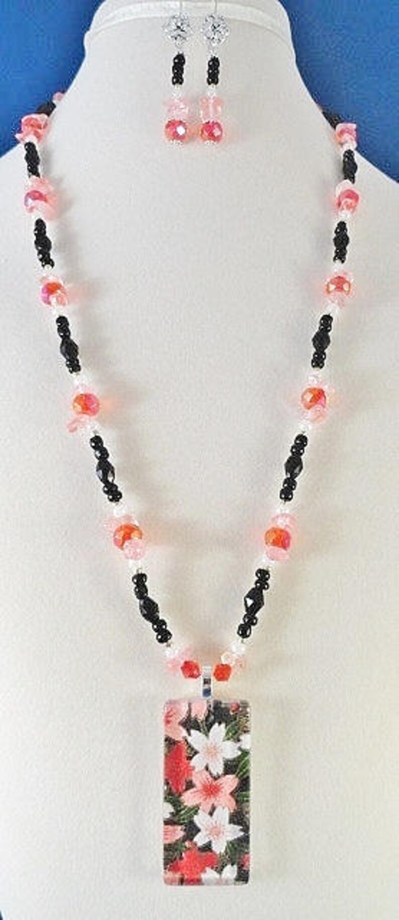 May include: A black and pink beaded necklace with a rectangular pendant featuring a floral design. The pendant is made of glass and has a pink, white, and green floral pattern.