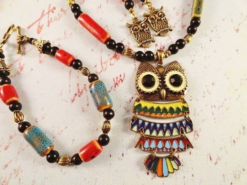 May include: A colorful owl pendant necklace with black and red beads. The owl has a yellow, green, blue, and orange design. The necklace comes with a matching bracelet and a pair of owl earrings.