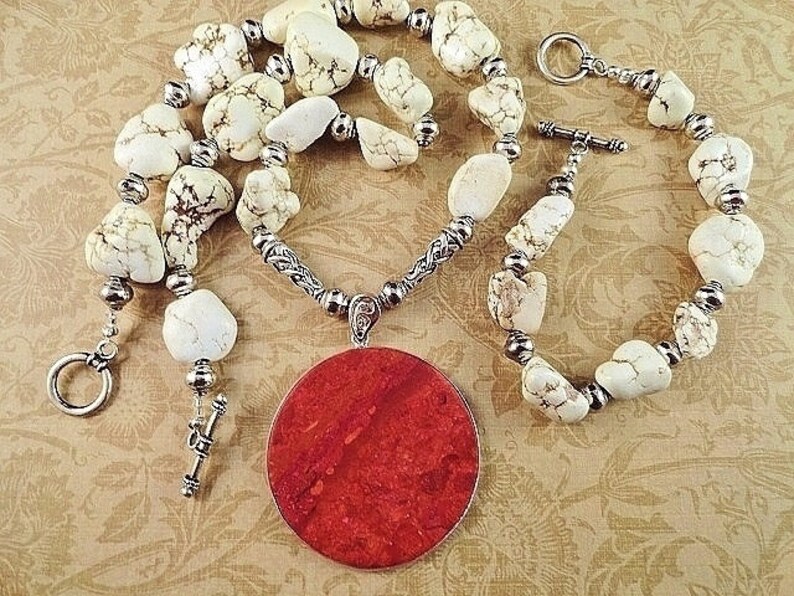 May include: A white and brown stone necklace with a large red pendant. The necklace has a silver chain and toggle clasp.
