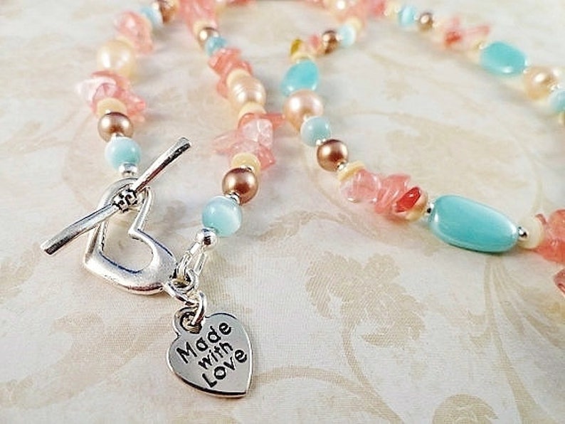 May include: A delicate necklace made with pastel pink, blue, and white beads. The necklace features a heart-shaped clasp with a charm that reads "Made with Love".