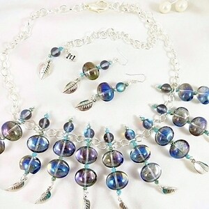 May include: A silver-toned necklace and earrings set featuring iridescent blue and purple glass beads. The necklace and earrings have a chain design with dangling beads and silver leaf charms. The jewelry set is displayed on a white surface.