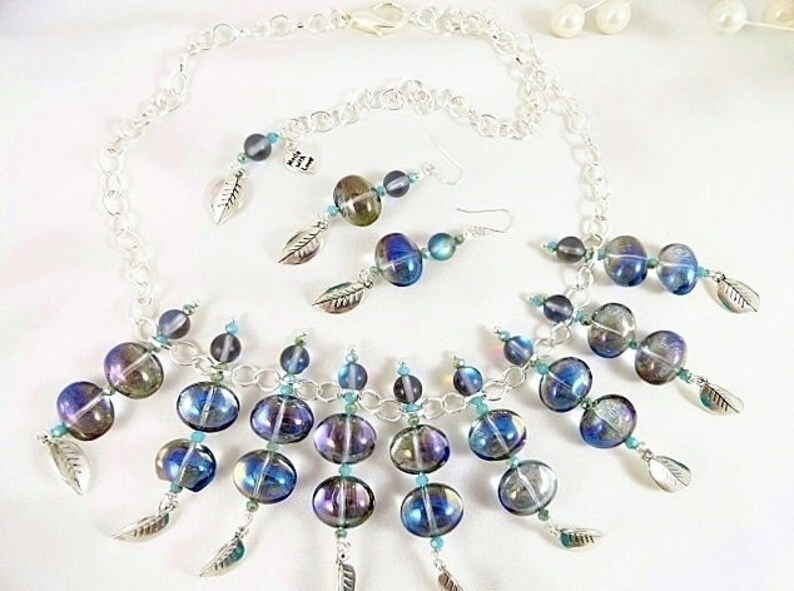 May include: A silver-toned necklace and earrings set featuring iridescent blue and purple glass beads. The necklace and earrings have a chain design with dangling beads and silver leaf charms. The jewelry set is displayed on a white surface.