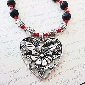May include: A silver heart-shaped pendant with a floral design hangs from a red and black beaded necklace.