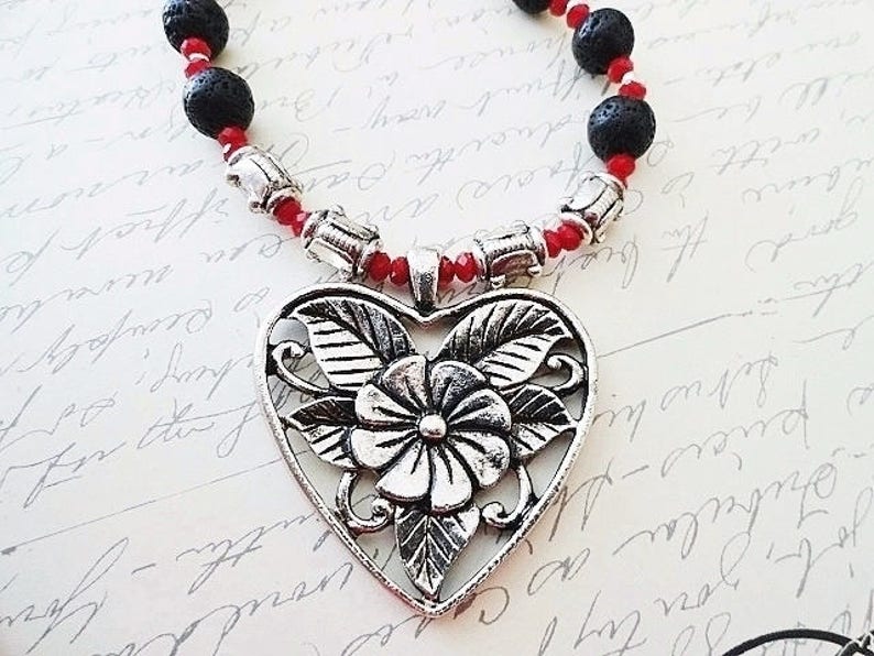 May include: A silver heart-shaped pendant with a floral design hangs from a red and black beaded necklace.