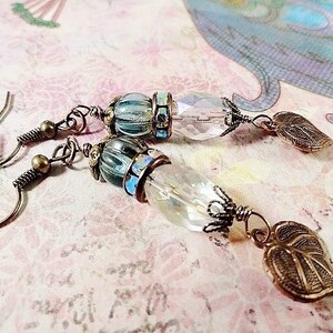 May include: A pair of earrings with a clear crystal bead, a blue and gold bead, and a leaf-shaped charm. The earrings are made with bronze wire and have a hook closure.
