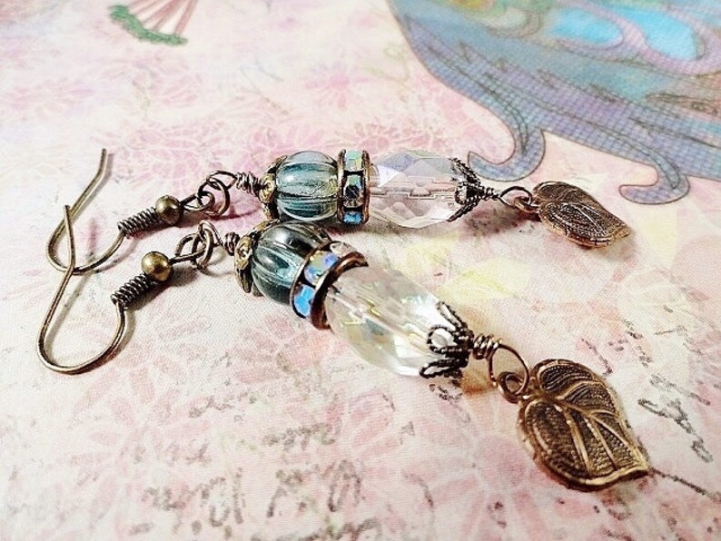 May include: A pair of earrings with a clear crystal bead, a blue and gold bead, and a leaf-shaped charm. The earrings are made with bronze wire and have a hook closure.