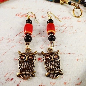 May include: A pair of owl charm earrings with red and black beads. The owls are gold-toned and have black eyes. The earrings are hanging from gold-toned hooks.