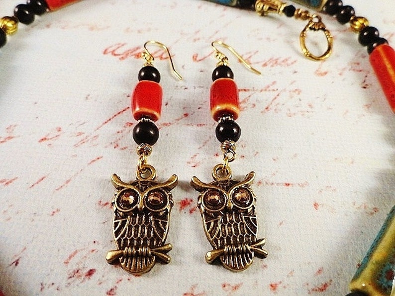 May include: A pair of owl charm earrings with red and black beads. The owls are gold-toned and have black eyes. The earrings are hanging from gold-toned hooks.