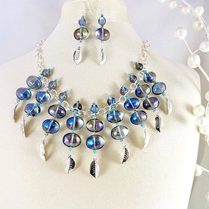 May include: A necklace and earring set featuring iridescent blue and purple glass beads. The necklace has a silver chain and dangling silver feather charms. The earrings mirror the necklace design. The jewelry is displayed on a mannequin.