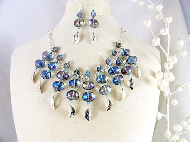 May include: A necklace and earring set featuring iridescent blue and purple glass beads. The necklace has a silver chain and dangling silver feather charms. The earrings mirror the necklace design. The jewelry is displayed on a mannequin.