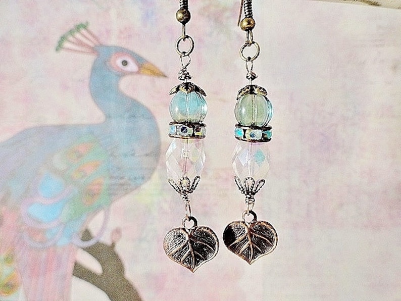 May include: A pair of dangle earrings with a blue and white glass bead, a clear crystal bead, and a leaf-shaped charm. The earrings are made with a bronze-colored metal.