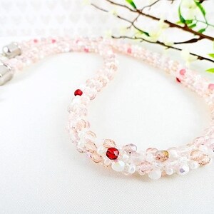 May include: A delicate necklace composed of small, light pink and clear beads, interspersed with a few red beads. The necklace has a silver clasp and is displayed on a white surface. The necklace is a piece of jewelry.