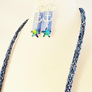 May include: A blue and silver necklace with a silver anchor charm and a blue and green star charm. The necklace is made of a braided cord.
