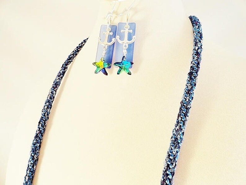 May include: A blue and silver necklace with a silver anchor charm and a blue and green star charm. The necklace is made of a braided cord.