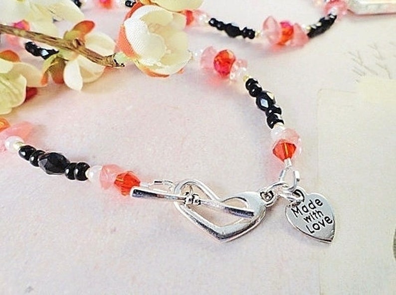 May include: A delicate beaded necklace with black, pink, and white beads. The necklace has a silver heart charm with the words "Made with Love" engraved on it.