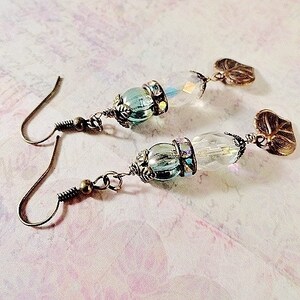 May include: A pair of earrings with a teardrop-shaped glass bead and a leaf-shaped charm. The beads are iridescent and have a light blue color. The charms are made of metal and have a bronze finish.