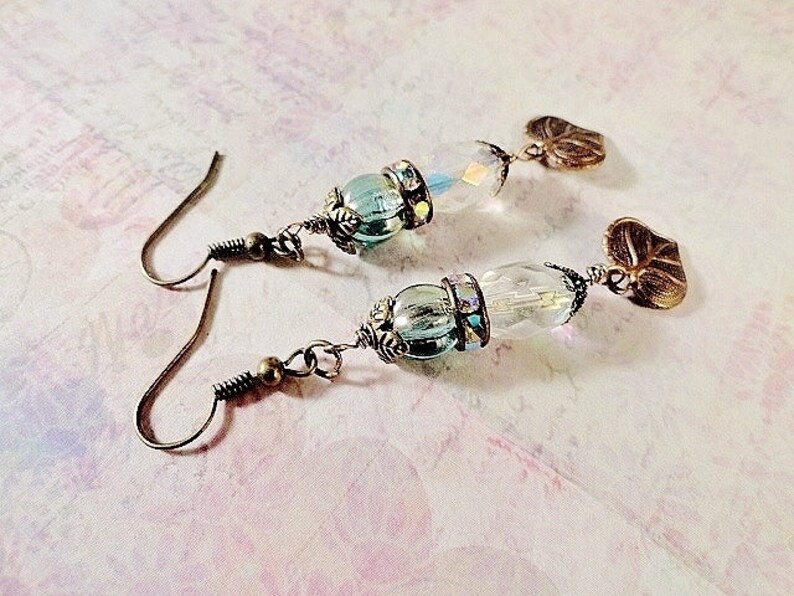 May include: A pair of earrings with a teardrop-shaped glass bead and a leaf-shaped charm. The beads are iridescent and have a light blue color. The charms are made of metal and have a bronze finish.