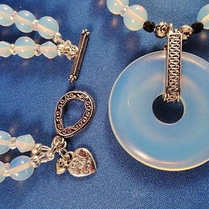May include: A multi-strand necklace with opal beads and silver accents. The necklace features a large, round opal pendant with a silver ring and a silver charm with the words "Peace Love & Joy".