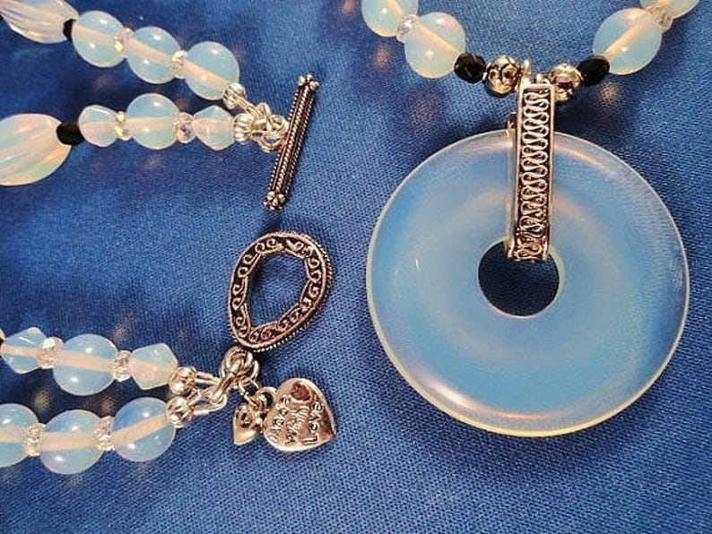 May include: A multi-strand necklace with opal beads and silver accents. The necklace features a large, round opal pendant with a silver ring and a silver charm with the words "Peace Love & Joy".