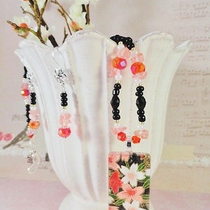 May include: A delicate necklace and earrings set featuring black beads, pink and clear glass beads, and a rectangular pendant with a floral design. The pendant has a black background with pink and white flowers.