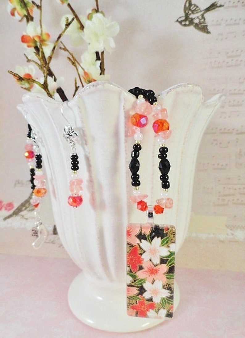 May include: A delicate necklace and earrings set featuring black beads, pink and clear glass beads, and a rectangular pendant with a floral design. The pendant has a black background with pink and white flowers.
