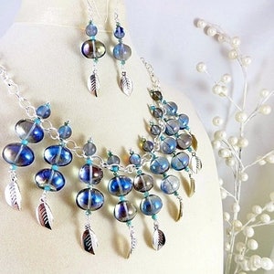 May include: A necklace and earring set featuring iridescent blue and silver glass beads. The necklace and earrings have silver leaf charms. The jewelry is displayed on a mannequin.