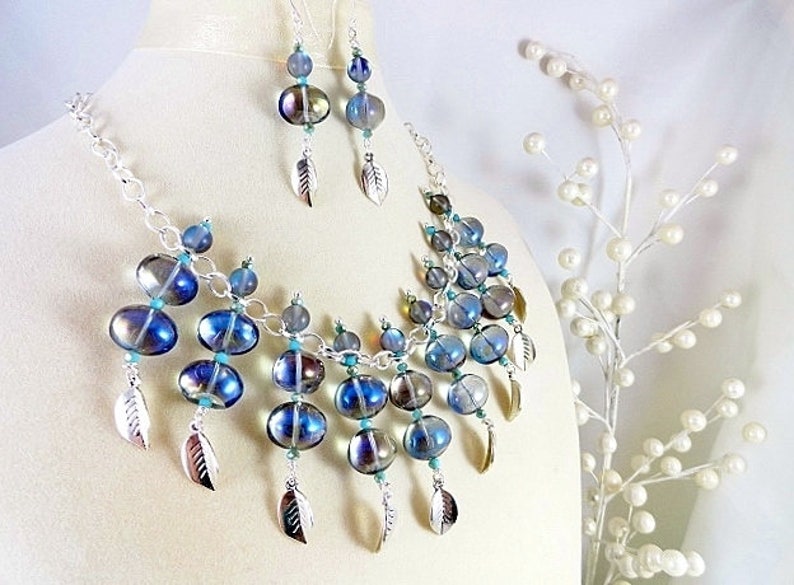 May include: A necklace and earring set featuring iridescent blue and silver glass beads. The necklace and earrings have silver leaf charms. The jewelry is displayed on a mannequin.