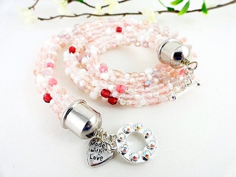 May include: A multi-strand beaded necklace in shades of pink, white, and red. The necklace features a silver clasp and a charm that says "Made with Love". The necklace is approximately 18 inches long.