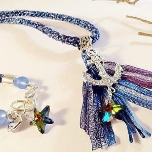 May include: A blue and purple braided cord necklace with a silver anchor charm and a blue and purple tassel. The tassel has a silver star charm.