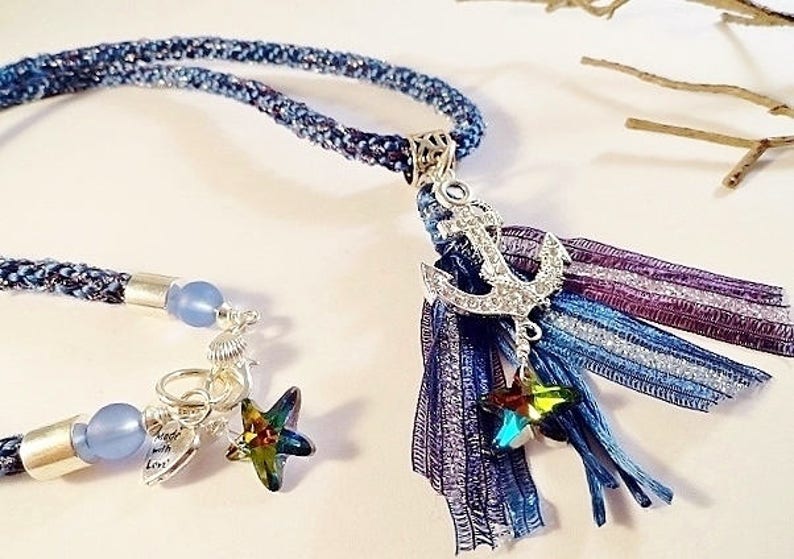 May include: A blue and purple braided cord necklace with a silver anchor charm and a blue and purple tassel. The tassel has a silver star charm.