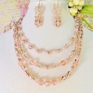 K&ouml;nnte beinhalten: Ein mehrreihiges Halsketten- und Ohrring-Set mit ros&eacute;goldenen und rosafarbenen Perlen. Die Halskette hat vier Reihen Perlen, und die Ohrringe sind im H&auml;nge-Stil mit einem herzf&ouml;rmigen Design.