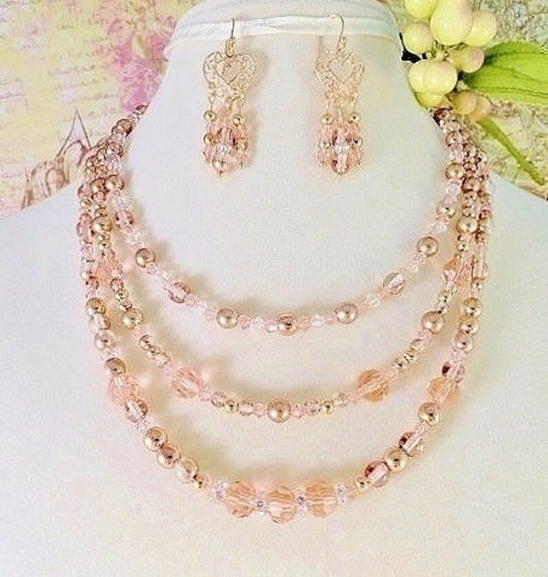 K&ouml;nnte beinhalten: Ein mehrreihiges Halsketten- und Ohrring-Set mit ros&eacute;goldenen und rosafarbenen Perlen. Die Halskette hat vier Reihen Perlen, und die Ohrringe sind im H&auml;nge-Stil mit einem herzf&ouml;rmigen Design.