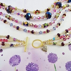 May include: Multi-strand necklace featuring gold beads and purple, pink, and clear crystals. The necklace has a gold toggle clasp and a small heart charm inscribed with the words "Made with Love".