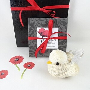 May include: A decorative bird figurine made of burlap with a yellow beak and gray wings. The image also shows a black gift box with a red ribbon, a smaller box with a poppy design, and red poppy cutouts. The text "Jewelry by PJ" is visible.