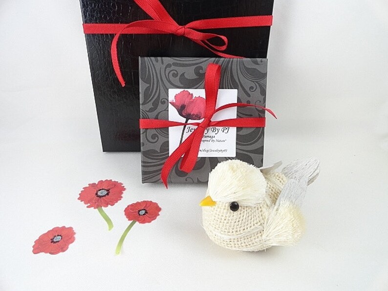 May include: A decorative bird figurine made of burlap with a yellow beak and gray wings. The image also shows a black gift box with a red ribbon, a smaller box with a poppy design, and red poppy cutouts. The text "Jewelry by PJ" is visible.