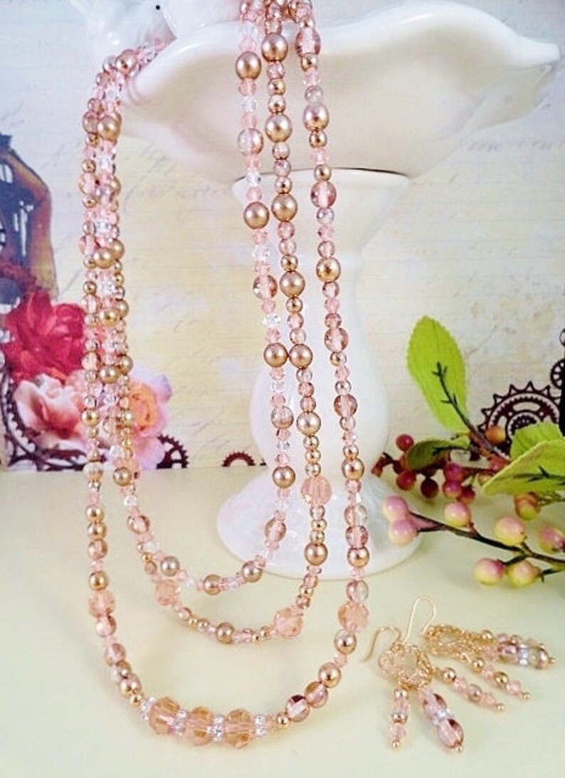 K&ouml;nnte beinhalten: Eine mehrreihige Halskette und ein passendes Ohrring-Set mit ros&eacute;goldenen und klaren Glasperlen. Die Halskette ist mit drei Perlenreihen geschichtet, und die Ohrringe sind im H&auml;nge-Stil mit mehreren Perlen.