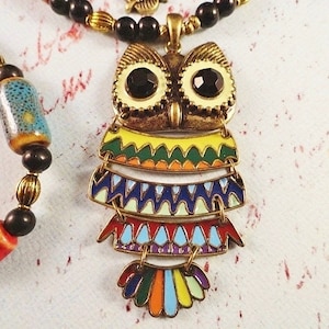 May include: A colorful owl pendant necklace with black beads and red and blue beads. The owl is made of metal and has a layered design with geometric patterns.